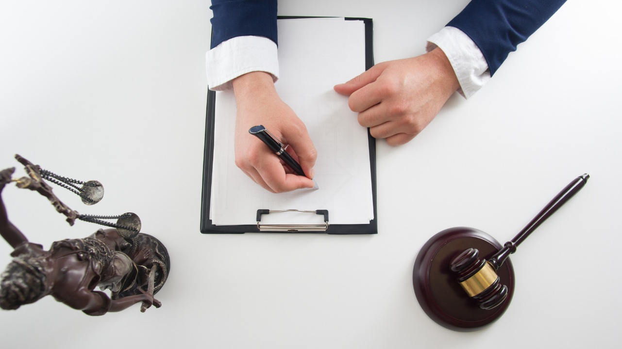 Image of a person writing on a clipboard with justice scale and gavel off to the side Image of a person writing on a clipboard with justice scale and gavel off to the side
